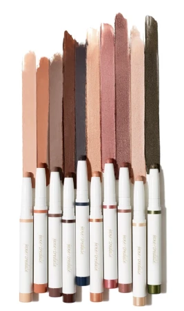 Na zdjęciu znajduje się zestaw kremowych cieni do powiek w sztyfcie marki Jane Iredale w różnych odcieniach. Widzimy 9 wysuniętych sztyftów z białymi opakowaniami i złotym napisem. Każdy cień ma inny kolor
