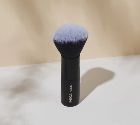 Na zdjęciu znajduje się pędzel do makijażu typu kabuki marki INIKA Organic. Pędzel stoi pionowo na jasnym tle, a jego czarny trzonek zakończony jest gęstym, zaokrąglonym włosiem, które u nasady jest ciemne, a na końcówkach przechodzi w jaśniejszy odcień