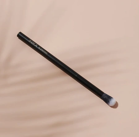 Na zdjęciu widoczny jest pędzel INIKA Organic Blending Brush, przeznaczony do blendowania cieni do powiek. Pędzel ma smukły, czarny trzonek z białym napisem i zaokrąglone, miękkie włosie w odcieniach bieli i czerni. Produkt jest na beżowym tle