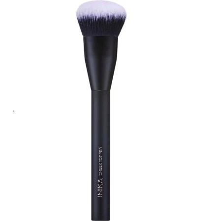 Na zdjęciu widoczny jest pędzel INIKA Organic Cheek Topper Brush. Ma gęste, miękkie, syntetyczne włosie o zaokrąglonym kształcie trzonek pędzla wykonany jest w eleganckim matowym, czarnym wykończeniu, z nazwą marki