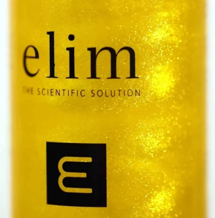 Zbliżenie etykiety kosmetyku Elim MEDIHEEL Rejuvenating Gold Spritz. Na tle złotego płynu z połyskującymi drobinkami widoczne czarne logo „elim – THE SCIENTIFIC SOLUTION” oraz symbol litery „E” w czarnej ramce.
