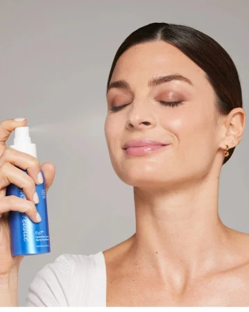 Na zdjęciu kobieta spryskuje twarz nawilżającą mgiełką D₂O™ Hydration Spray od Jane Iredale. Ma zamknięte oczy i zrelaksowany wyraz twarzy. Produkt widoczny w jej dłoni to niebieska butelka z białym atomizerem – idealny do skóry suchej.