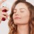 Na zdjęciu kobieta spryskuje twarz nawilżającą mgiełką Pommist Hydration Spray od Jane Iredale. Ma zamknięte oczy i zrelaksowany wyraz twarzy. Produkt widoczny w jej dłoni to czerwona butelka z białym atomizerem – idealny dla każdego rodzaju skóry.