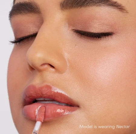Na zdjęciu widzimy modelkę aplikującą błyszczyk do ust Jane Iredale w odcieniu Nectar. Usta modelki mają subtelny, brzoskwiniowo-beżowy odcień z połyskiem, który dodaje świeżości i naturalnego blasku.