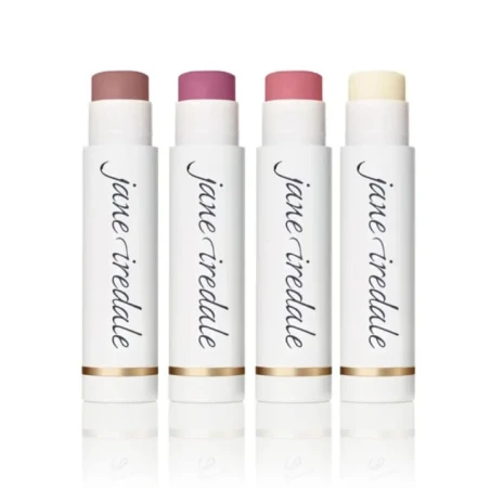 Na zdjęciu widoczne są cztery sztyfty pomadek ochronnych Jane Iredale. Produkty te mają eleganckie, białe opakowania z logo marki i złotym akcentem przy podstawie. Każdy ma inny odcień – od neutralnych beży i różów po klasyczny bezbarwny balsam.