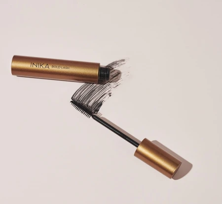 Na zdjęciu widoczny jest tusz do rzęs INIKA Organic Bold Lash Mascara w eleganckim, metaliczno-złotym opakowaniu. Maskara została otwarta, a szczoteczka z produktem pozostawiła wyrazisty, czarny ślad tuszu na jasnym tle.