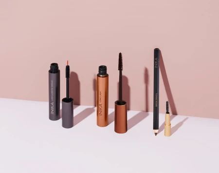 Na zdjęciu widoczne są cztery kosmetyki marki INIKA Organic ustawione na jasnoróżowym tle. Od lewej: Eyeliner w płynie, Tusz do rzęs Bold Lash Mascara, Kredka do brwi lub oczu