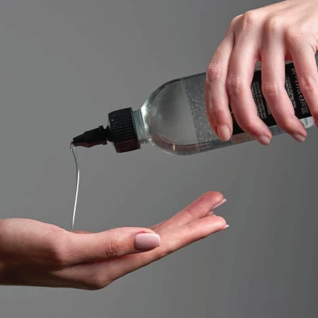Osoba wyciska Davines OI Liquid Luster z przezroczystej butelki na dłoń. Przezroczysty płyn wypływa przez aplikator, a tło jest jednolite i szare. Produkt stosowany do pielęgnacji i nabłyszczania włosów.