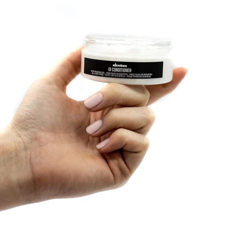 Dłoń trzymająca słoiczek odżywki do włosów Davines OI Conditioner. Pojemnik jest biały z czarną etykietą, bez zakrętki, ukazujący kremową konsystencję produktu w środku.