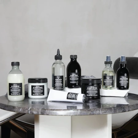 Zestaw produktów do pielęgnacji włosów marki Davines z linii OI ustawiony na marmurowym stoliku; wśród nich szampon, odżywka, olejek, mleczko, masełko i lotion, wszystkie w charakterystycznych czarno-białych opakowaniach.