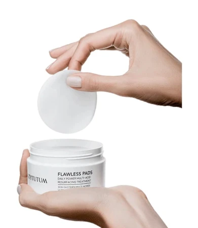 Na zdjęciu widoczne są dłonie osoby sięgającej po pojedynczy płatek kosmetyczny z opakowania Flawless Pads marki Instytutum. Produkt to codzienny zabieg złuszczający z kwasami, który poprawia strukturę i wygląd skóry.