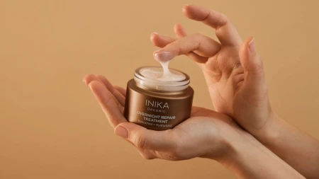 Na zdjęciu przedstawiono słoiczek INIKA Organic Overnight Repair Treatment trzymany w dłoniach. Jedna z dłoni delikatnie nabiera kremową formułę produktu, co podkreśla jego lekką, jedwabistą konsystencję.