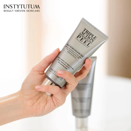 Dłoń trzyma srebrną tubkę Instytutum Triple Action Resurfacing Peel. W tle rozmyta druga tubka tego samego produktu. Peeling złuszczający z kwasami i enzymami poprawia strukturę skóry. W lewym górnym rogu logo Instytutum.