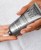 Dłoń wyciska biały peeling z tubki Instytutum Triple Action Resurfacing Peel na wnętrze drugiej dłoni. Tubka srebrna z czarnym napisem, tło stanowi biały ręcznik. Produkt złuszcza, wygładza i nawilża skórę. Paznokcie zadbane, w jasnym odcieniu.