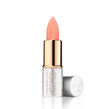 Na zdjęciu widoczna jest pomadka Just Kissed Lip & Cheek Stain marki Jane Iredale. Produkt ma eleganckie opakowanie w odcieniach srebra i złota, z odkrytą szminką w brzoskwiniowym kolorze.  Jest to wielofunkcyjny kosmetyk do ust i policzków