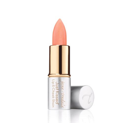 Na zdjęciu widoczna jest pomadka Just Kissed Lip & Cheek Stain marki Jane Iredale. Produkt ma eleganckie opakowanie w odcieniach srebra i złota, z odkrytą szminką w brzoskwiniowym kolorze.  Jest to wielofunkcyjny kosmetyk do ust i policzków