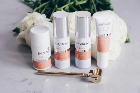 Zdjęcie wszystkich produktów z linii Environ Focus Care Radiance, stojących na tle kwiatów, na marmurowej powierzchni. Przed produktami leży złoty Roll Cit.