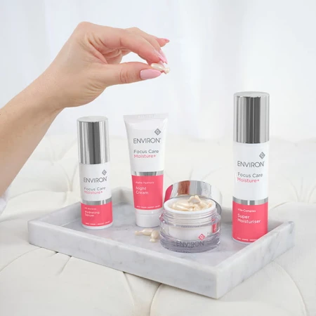 Wszystkie produkty linii Environ Moisture, stojące obok siebie na marmurkowej tacy, jasnym tle. Damskie dłoń trzyma nad produktami jedną kapsułkę nawilżającą.