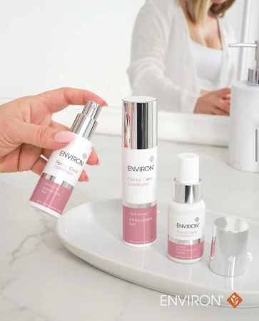 Na dekoracyjnej białej podstawce, kobieca dłoń trzyma ENVIRON Colostrum Gel w otoczeniu innych produktów z linii Environ Focus Care Comfort+