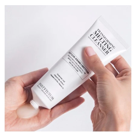 Dwie dłonie trzymają tubkę Instytutum Transforming Melting Cleanser. Jedna wyciska produkt na dłoń. Tubka biała z czarnym napisem, zawiera oczyszczający i nawilżający żel z kwasem hialuronowym, olejem migdałowym i witaminami.