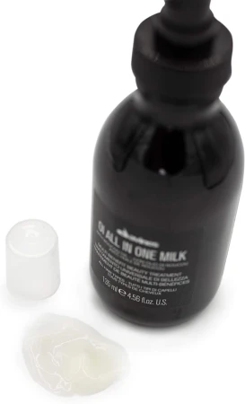 Butelka Davines OI All In One Milk o pojemności 135 ml z przezroczystą nakrętką obok. Przed butelką widoczna próbka mleczka o kremowej konsystencji, rozprowadzona na białej powierzchni.
