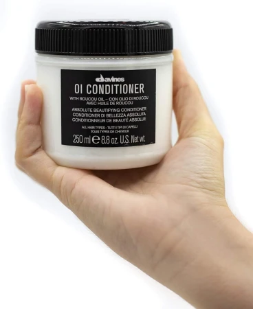 Osoba trzyma w dłoni słoiczek odżywki Davines OI Conditioner 250 ml z czarną nakrętką i czarną etykietą. Na etykiecie znajdują się informacje o zawartości oleju roucou i przeznaczeniu dla wszystkich typów włosów.