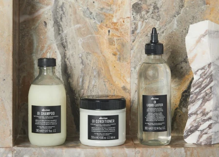 Trzy kosmetyki do włosów marki Davines ustawione na marmurowej półce: OI Shampoo, OI Conditioner oraz Liquid Luster – każdy w charakterystycznym opakowaniu z czarną etykietą, na tle kamiennej ściany o naturalnym wzorze.