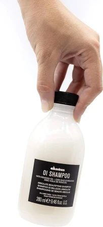 Ręka trzymająca butelkę szamponu Davines OI Shampoo o pojemności 280 ml; butelka z białym płynem, czarną zakrętką i czarną etykietą z białym tekstem.