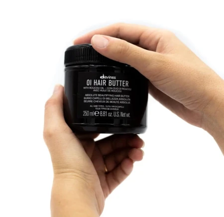 Ręce trzymające czarny słoik kosmetyku Davines OI Hair Butter o pojemności 250 ml. Produkt przeznaczony do pielęgnacji włosów, etykieta w kolorze czarnym z białym napisem. Tło białe.