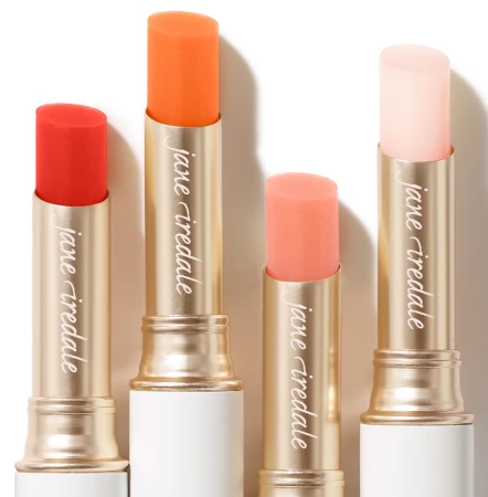 Na zdjęciu cztery pomadki ochronne marki Jane Iredale w sztyfcie. Każda z nich ma inny odcień: intensywnie czerwony, energetyczny pomarańczowy, delikatny brzoskwiniowy i przezroczysty różowy. Opakowania są eleganckie, złoto białe, z połyskiem.
