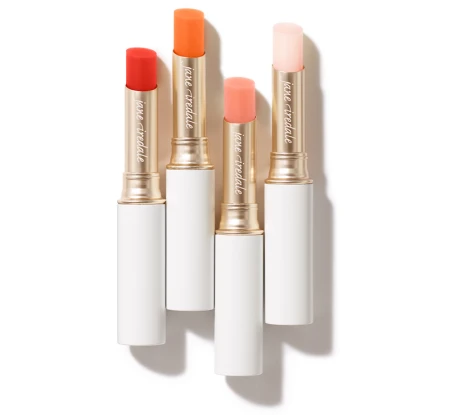 Na zdjęciu widoczne są cztery pomadki z kolekcji Just Kissed Lip and Cheek Stain marki Jane Iredale. Produkty umieszczone są w eleganckich, białych opakowaniach ze złotym wykończeniem.  Każda pomadka ma inny odcień