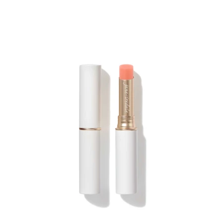 Na zdjęciu znajduje się elegancka pomadka ochronna marki Jane Iredale. Produkt ma biało-złote opakowanie o minimalistycznym i luksusowym wyglądzie. Pomadka jest wysunięta — ma brzoskwiniowo-różowy odcień