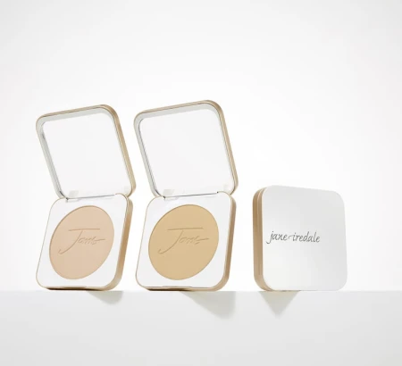 Na zdjęciu są kompaktowe opakowania wielokrotnego użytku marki jane iredale.  Mminimalistyczny, elegancki design w kolorze złotym i białym,  napis na froncie: “you make the world more beautiful. Wewnątrz puder.