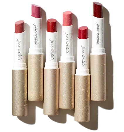 Na zdjęciu znajduje się kolekcja szminek Jane Iredale z serii Color Luxe. Produkty są umieszczone w eleganckich, złotych opakowaniach z subtelnym połyskiem . Widać krople wody na sztyftach. Grafika jest świeża i soczysta,