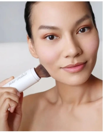 Na zdjęciu widać kobietę aplikującą puder za pomocą pędzla jane iredale Powder-Me SPF. Produkt ma formę wygodnego aplikatora z pędzlem i jest używany na zewnątrz, w słoneczny dzień, co podkreśla jego funkcję ochrony przeciwsłonecznej (SPF).