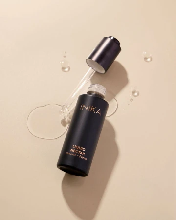 Na zdjęciu widoczny jest otwarty kosmetyk INIKA Organic Liquid Nectar – Nourish + Prime. Buteleczka w eleganckim, matowym, ciemnogranatowym odcieniu z subtelnym złotym napisem leży na jasnym tle. Obok niej znajduje się zakrętka z pipetą
