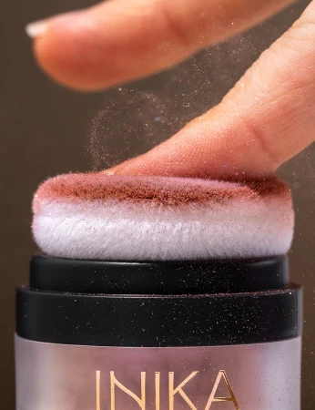 Na zdjęciu widoczny jest produkt INIKA Organic Puff Pot – róż do policzków z aplikatorem typu puff.  Zbliżenie ukazuje gąbeczkę nasączoną różowym pudrem, delikatnie muskającą opuszek palca
