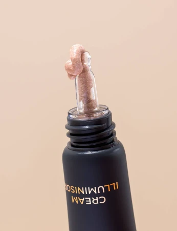 Na zdjęciu znajduje się tubka kosmetyku w ciemnym, matowym opakowaniu z napisem „CREAM ILLUMINISOR”. Z tubki wystaje przezroczysty aplikator, na którego końcówce widoczna jest kremowa, błyszcząca substancja o różowo-złotym odcieniu.