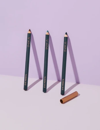 Na zdjęciu widoczne są trzy kredki do oczu ustawione pionowo na jasnym tle z lekkim fioletowym odcieniem. Każda kredka ma ciemny korpus z napisem „INIKA EYE PENCIL”, a ich końcówki różnią się kolorami. Obok leży złota skuwka
