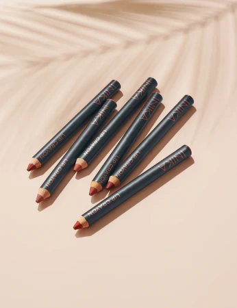 Na zdjęciu znajduje się sześć kredek do ust INIKA Organic Lip Crayon, rozmieszczonych ukośnie na tle w ciepłym odcieniu nude z delikatnym cieniem liścia palmy, co nadaje kompozycji letniego, naturalnego klimatu.