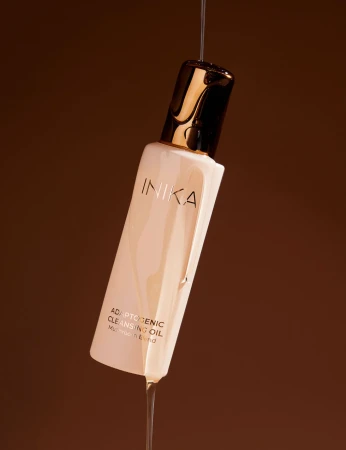Zdjęcie ukazuje INIKA Adaptogenic Cleansing Oil w eleganckiej kompozycji z dynamicznie spływającą strugą olejku, która oplata butelkę.  Ciemne, ciepłe tło podkreśla luksusowy charakter produktu i kontrastuje z jasnym opakowaniem oraz złotą nakrętką.