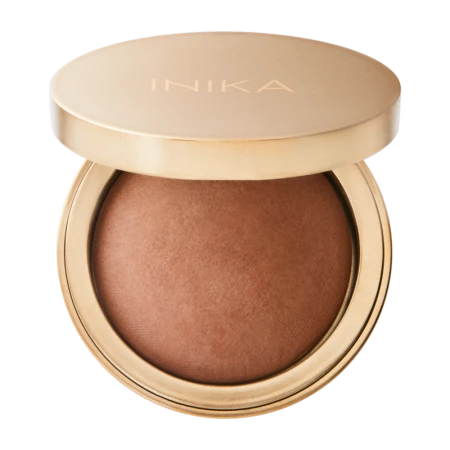 Na zdjęciu widoczny jest prasowany bronzer marki INIKA Organic w eleganckim, okrągłym opakowaniu w kolorze satynowego złota. Pokrywka z subtelnym logo "INIKA" została lekko uchylona, ukazując wypiekany kosmetyk o ciepłym, karmelowo-brązowym odcieniu