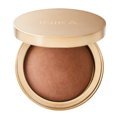 Na zdjęciu widoczny jest prasowany bronzer marki INIKA Organic w eleganckim, okrągłym opakowaniu w kolorze satynowego złota. Pokrywka z subtelnym logo "INIKA" została lekko uchylona, ukazując wypiekany kosmetyk o ciepłym, karmelowo-brązowym odcieniu