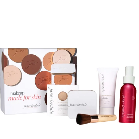 Na zdjęciu widzimy elegancki, zestaw kosmetyków w przy białym pudełku marki jane iredale. Widzimy puder, puderniczkę, spray, bazę i pędzel handi.