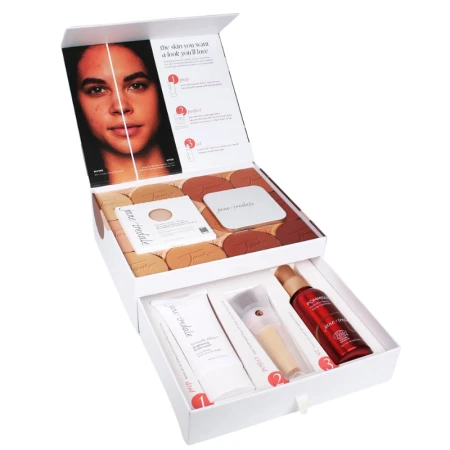Na zdjęciu widzimy elegancki, zestaw kosmetyków w otwartym, białym pudełku marki jane iredale. Widzimy puder, puderniczkę, spray, bazę i pędzel handi.