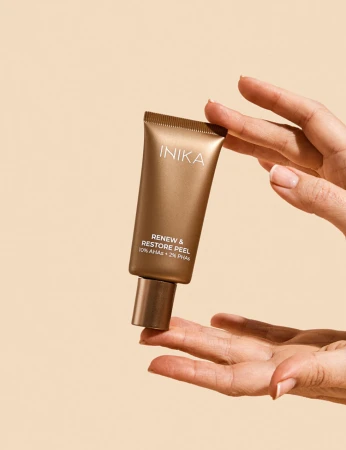 Na zdjęciu przedstawiono produkt marki INIKA Organic – Renew & Restore Peel (peeling odnawiający), trzymany delikatnie w powietrzu pomiędzy dłońmi. Elegancka, brązowo-złota tuba z minimalistycznym napisem podkreśla luksusowy charakter kosmetyku.
