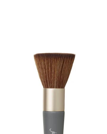 Na zdjęciu zbliżenie na główkę pędzla do makijażu marki Jane Iredale. Kształt główki: płaska, szeroka i gęsto zbita. Włosie: syntetyczne, miękkie, o brązowym kolorze. Trzonek: krótki, w kolorze szaro-grafitowym z logiem „jane iredale”