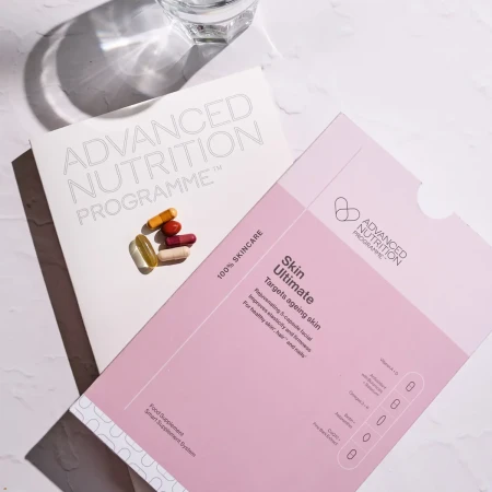 zestaw suplementów diety Skin Ultimate marki Advanced Nutrition Programme – różowe opakowanie z opisem i kapsułkami, obok biała teczka z logo programu oraz szklanka wody.