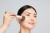 Na zdjęciu widzimy kobietę, której twarz jest pokrywana pudrem przy użyciu pędzla Jane Iredale Multiuse. Pędzel ma elegancki wygląd — jasne włosie przechodzące w ciemniejsze u nasady, złotą skuwkę oraz szarą rączkę z logotypem.