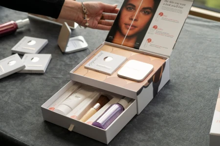 Na zdjęciu widzimy elegancki, zestaw kosmetyków w przy białym pudełku marki jane iredale. Widzimy puder, puderniczkę, spray, bazę i pędzel handi.